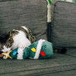 Load image into Gallery viewer, A tabby and white cat lies on a dark gray sofa, playfully biting and pawing at a Feline Frenzy O Christmas Tree kicker plush toy with white trim and colorful lights. A peach pillow is visible on the left.
