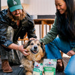 Load image into Gallery viewer, Dog parents petting their dog with a Cabot Creamery x Play Vermont Cheddar Shredder dog toy and the actual Cabot Creamery shredded cheese bag next to each other on a wooden deck.
