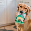 Load image into Gallery viewer, Dog holding a Cabot Creamery x Play Vermont Cheddar Shredder dog toy inspired by a bag of shredded cheese in its mouth inside a kitchen.
