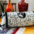 Load image into Gallery viewer, A black-and-white cat walking through a Feline Frenzy Purr &amp; Pounce Cat Tunnel toy in Black Celestial print, poking its head out of the peephole, set on a patterned rug in a living room with wooden floors and decorative vases.
