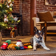 Load image into Gallery viewer, A small black, white, and tan dog lies on a woven rug next to a decorated Christmas tree with the Play Merry Minis Collection consisting of several colorful plush toys in a cozy living room with brick walls and a sofa in the background.
