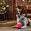 Load image into Gallery viewer, A small Australian Shepherd dog lies on a woven rug in a cozy living room, holding a Play Merry Minis Holiday Hauler plush red vintage truck toy with green trim. A decorated Christmas tree and brick fireplace are visible in the background.
