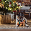 Load image into Gallery viewer, A small black, white, and tan dog lies on a woven rug in front of a decorated Christmas tree, holding a Play Merry Minis Holly Jolly Gingerbread Man cookie toy.
