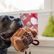 Load image into Gallery viewer, P.L.A.Y. Home for the Holidays Fur-Ever Naughty Toy - in mouth of big black dog looking to the side in front of a white bench with a gift bag and Christmas Tree pillows
