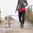 Load image into Gallery viewer, P.L.A.Y.'s Explorer Pack in Lava Red being worn around the waist of a woman holding her coffee and using hands-free leash feature while on a walk with her dog
